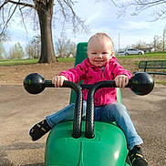 Chevelle is registered to the contest to win money with this photo: bench, child, daylight, fun, green_toy, grip, happy, jeans, outdoor, park, person, pink_jacket, play_equipment, playground, puma, smiling, sneakers, toddler, toy_vehicle, tree