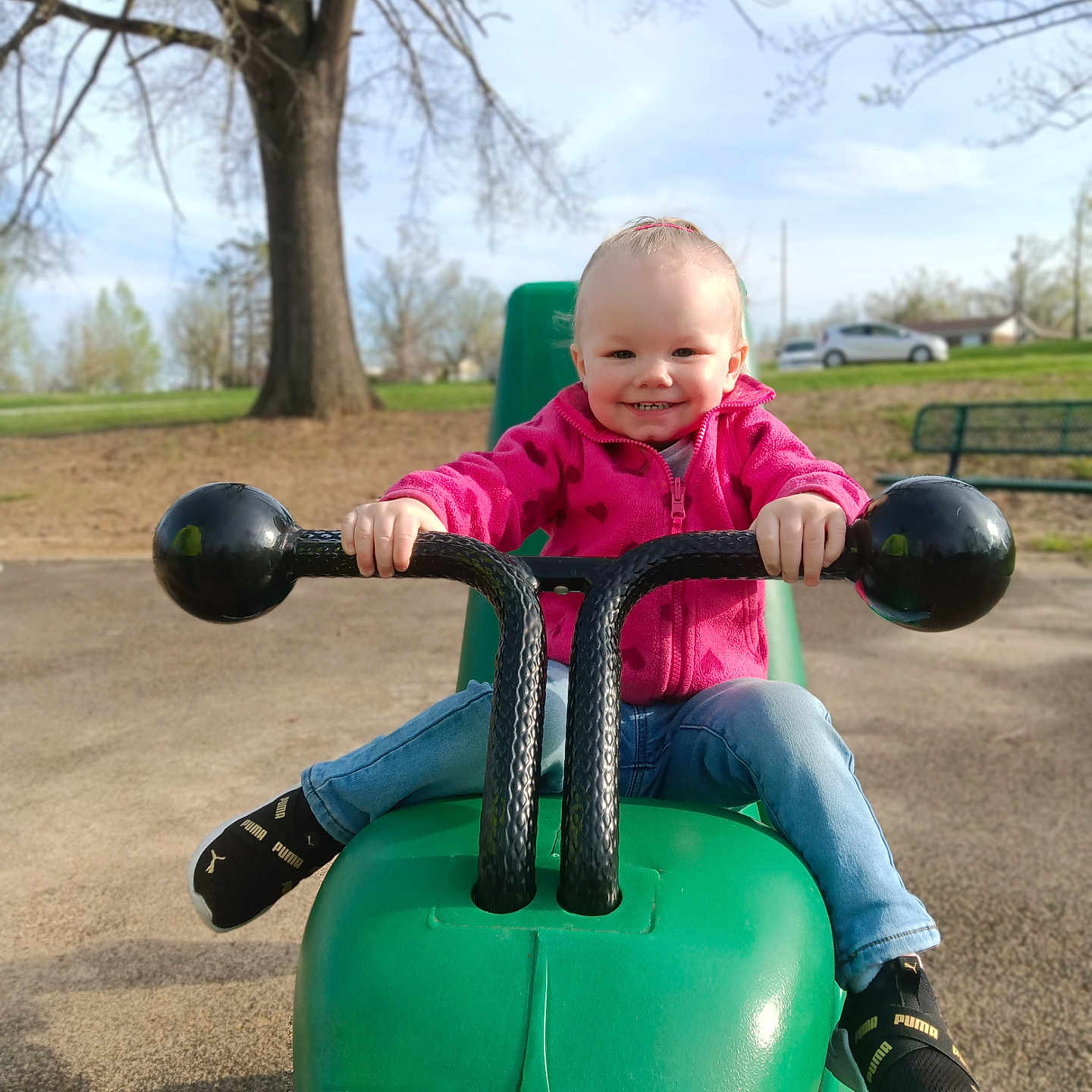 Chevelle is registered to the contest to win money with this photo: bench, child, daylight, fun, green_toy, grip, happy, jeans, outdoor, park, person, pink_jacket, play_equipment, playground, puma, smiling, sneakers, toddler, toy_vehicle, tree