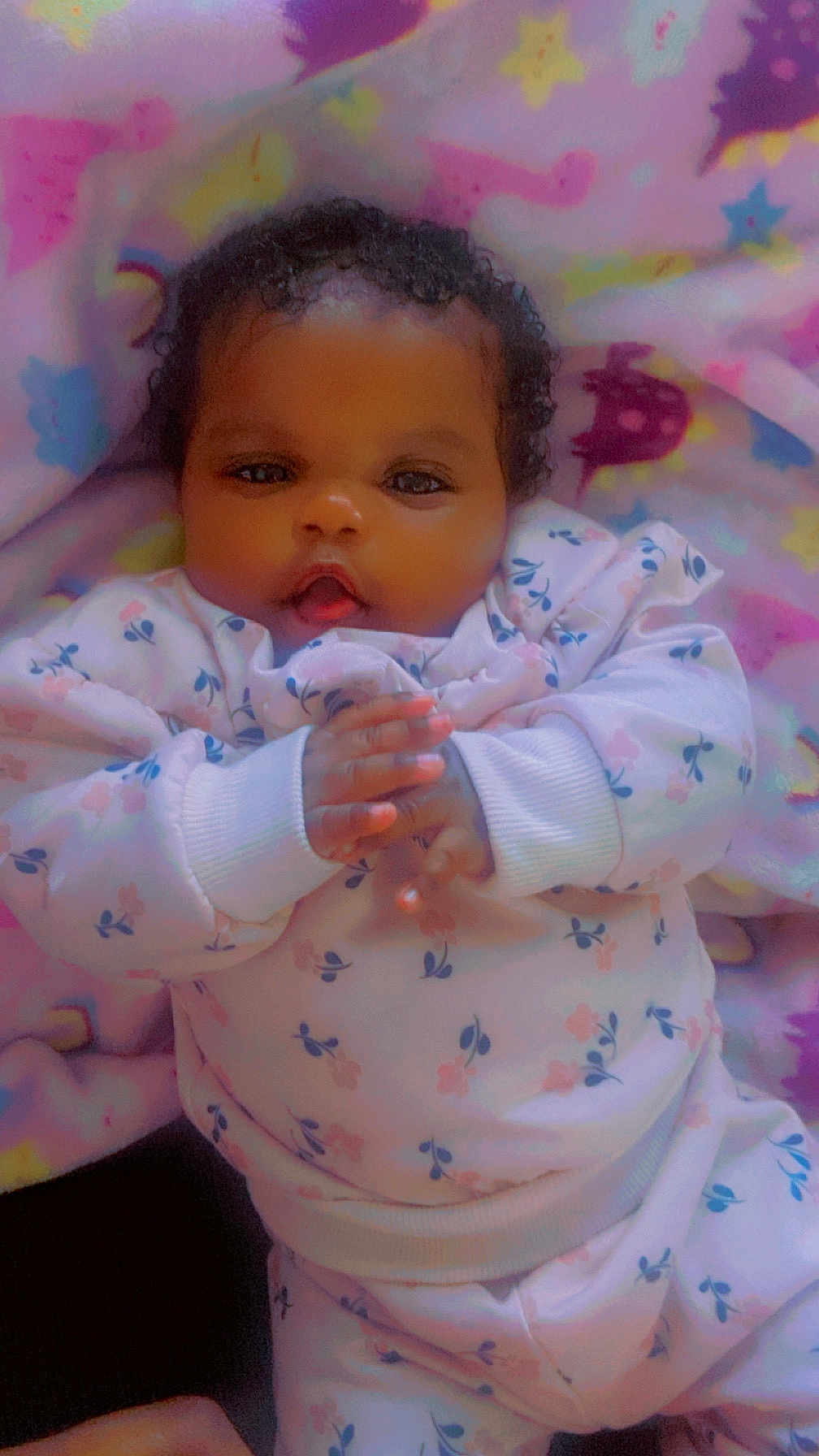 Deasia is registered to the contest to win money with this photo: baby, infant, child, curly_hair, blanket, floral_outfit, hands, clothing, cute, lying_down, portrait, face, eyes, indoor, soft_lighting, pink, blue, yellow, star_pattern, warm