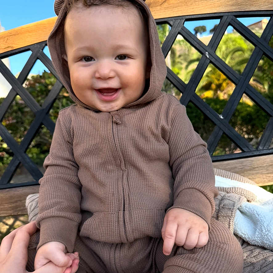 Malakhi joined the competition — help win amazing prizes! baby, child, smiling, hoodie, brown_clothing, outdoor, bench, hand_holding, portrait, happy, face, skin, person, blue_sky, greenery, daylight, photography, infant, cute, onesie