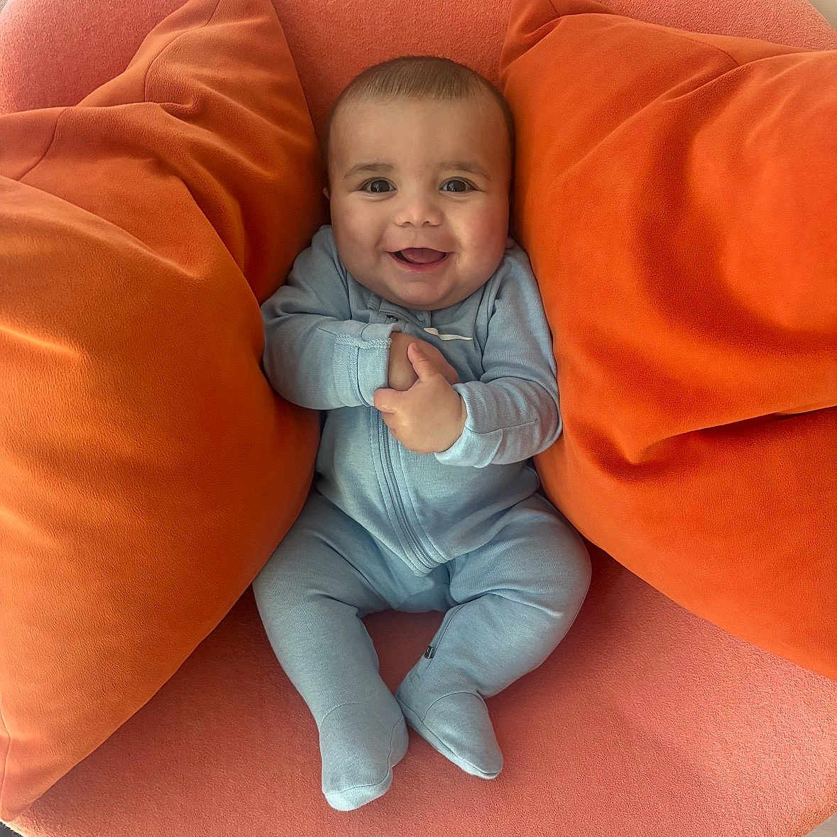 Mateo is registered to the contest to win money with this photo: baby, smiling, blue_onesie, orange_chair, orange_pillows, infant, happy, cute, child, indoor, soft_furniture, plush, seated, hands_clasped, portrait, cozy, person, head, feet, light