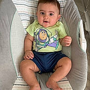 Jesse is registered to the contest to win money with this photo: baby, child, infant, baby_seat, buzz_lightyear, tshirt, shorts, barefoot, indoors, flooring, wood_floor, patterned_fabric, cute, smiling, sitting, person, face, young_child, happy, comfortable