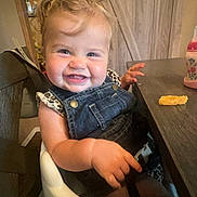 Charlie is registered to the contest to win money with this photo: toddler, child, smiling, high_chair, denim_overalls, leopard_print, table, cheese_puff, indoor, wooden_furniture, happy, person, face, baby_food, feeding_time, hair, skin, seatbelt, cheeks, cute