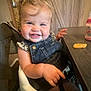 toddler, child, smiling, high_chair, denim_overalls, leopard_print, table, cheese_puff, indoor, wooden_furniture, happy, person, face, baby_food, feeding_time, hair, skin, seatbelt, cheeks, cute
