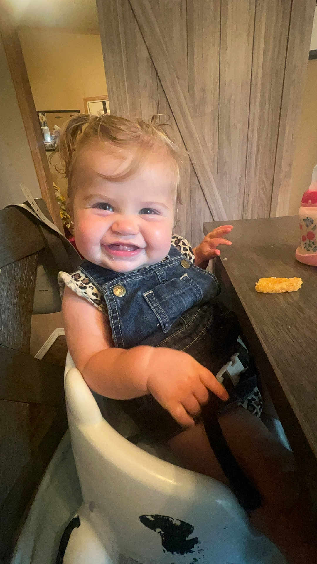 Charlie is registered to the contest to win money with this photo: toddler, child, smiling, high_chair, denim_overalls, leopard_print, table, cheese_puff, indoor, wooden_furniture, happy, person, face, baby_food, feeding_time, hair, skin, seatbelt, cheeks, cute
