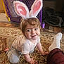 toddler, bunny_ears, child, crawling, carpet, indoor, playful, smiling, baby_gate, blanket, foot, sock, pants, person, headband, pink, white, brown_hair, striped_shirt, gray_pants