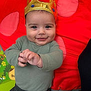 Kash is registered to the contest to win money with this photo: baby, crown, smiling, infant, play_mat, red_blanket, polka_dots, child, cute, indoors, portrait, happy, clothing, green_shirt, gray_pants, hand_gesture, face, headwear, young_child, playtime
