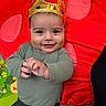 baby, crown, smiling, infant, play_mat, red_blanket, polka_dots, child, cute, indoors, portrait, happy, clothing, green_shirt, gray_pants, hand_gesture, face, headwear, young_child, playtime