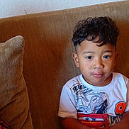 Danny Danio Mackos is registered to the contest to win money with this photo: child, toddler, portrait, sitting, couch, sofa, cushion, curly_hair, short_hair, smiling, face, eyes, white_shirt, graphic_shirt, snack, indoors, home, furniture, cozy, photography