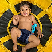 Fabian is registered to the contest to win money with this photo: toddler, child, smiling, shirtless, shorts, toy, water_gun, swing, yellow, black, grass, outdoor, play, happy, barefoot, dirt, fun, summer, face, person