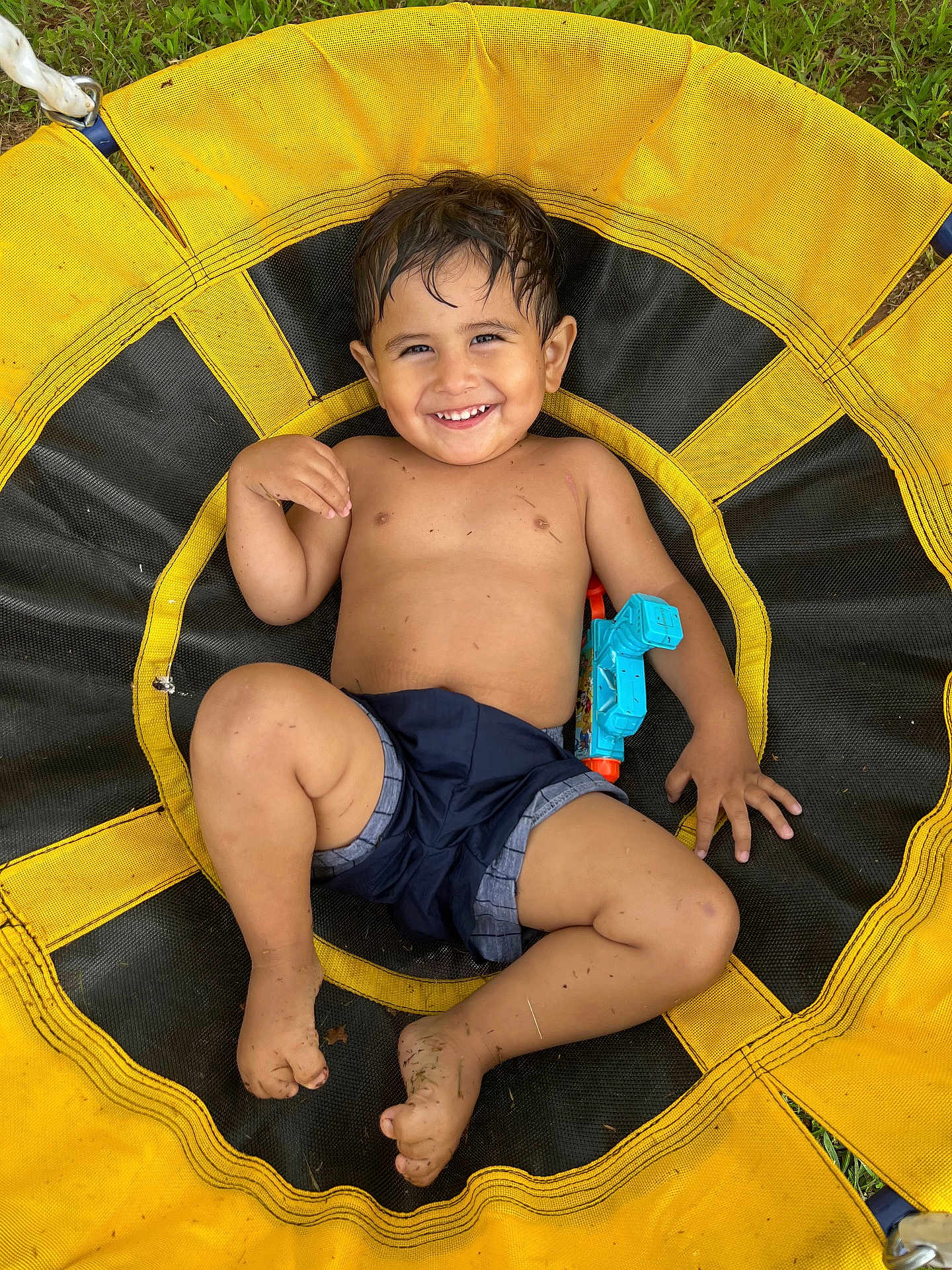 Fabian is registered to the contest to win money with this photo: toddler, child, smiling, shirtless, shorts, toy, water_gun, swing, yellow, black, grass, outdoor, play, happy, barefoot, dirt, fun, summer, face, person