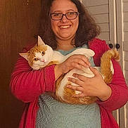 Hercules is registered to the contest to win money with this photo: person, cat, glasses, red_cardigan, orange_cat, white_cat, indoor, smiling, holding, pet, furniture, door, closet, happy, curly_hair, casual_clothing, portrait, cozy, warm_lighting, relaxed