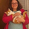 person, cat, glasses, red_cardigan, orange_cat, white_cat, indoor, smiling, holding, pet, furniture, door, closet, happy, curly_hair, casual_clothing, portrait, cozy, warm_lighting, relaxed