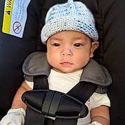 Leon is registered to the contest to win money with this photo: baby, infant, car_seat, knit_hat, mittens, safety_harness, warning_label, white_clothing, black_straps, closeup, face, serious_expression, child, seatbelt, indoor, soft_lighting, cute, newborn, portrait, seated