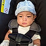 baby, infant, car_seat, knit_hat, mittens, safety_harness, warning_label, white_clothing, black_straps, closeup, face, serious_expression, child, seatbelt, indoor, soft_lighting, cute, newborn, portrait, seated
