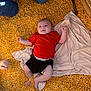 baby, black_shorts, blanket, blue_jeans, casual, cereal, clothing, corn_kernels, daytime, hands, infant, legs, lying_down, outdoor, person, red_shirt, relaxing, skin, sunlight, texture
