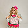 child, girl, curly_hair, pink_bow, watermelon_dress, white_chair, sitting, smiling, barefoot, studio, portrait, cute, happy, fashion, colorful_clothing, young_child, indoor, light_background, playful, innocence