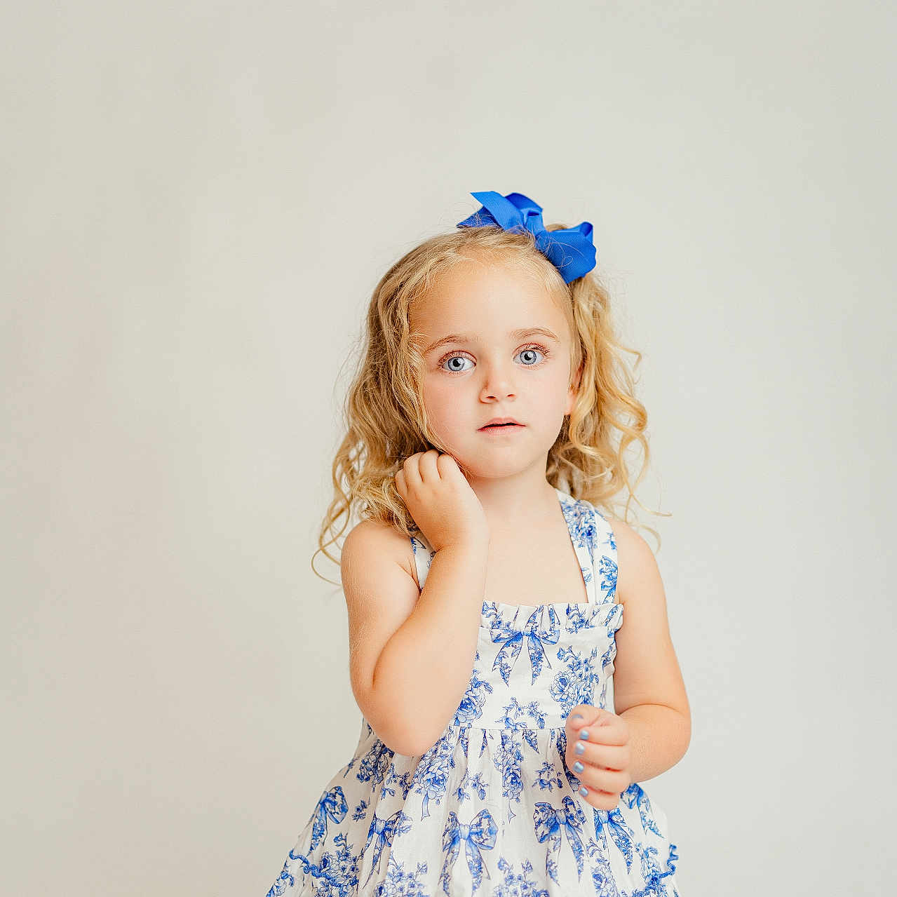 Brynlee is registered to the contest to win money with this photo: child, girl, blue_bow, curly_hair, blonde_hair, blue_eyes, dress, floral_pattern, white_dress, standing, portrait, studio_lighting, innocent, expression, cute, fashion, youth, person, background, photography