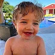 Raylynn is registered to the contest to win money with this photo: toddler, child, smiling, wet_hair, outdoor, poolside, blue_sky, sunny, happy, face, skin, eyes, mouth, teeth, snack, wrapper, background, red_building, green_chair, furniture