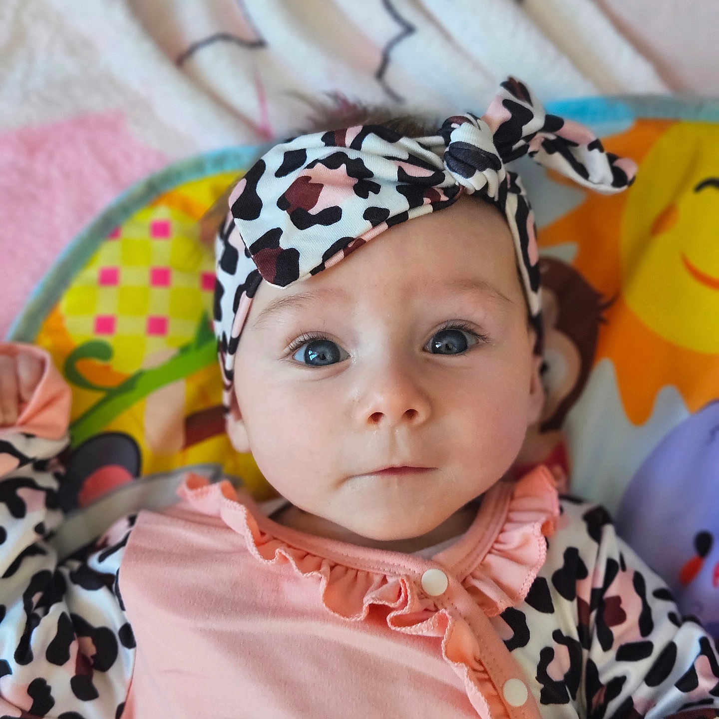 Lola participe au concours pour gagner de l'argent avec cette photo : baby, infant, headband, blue_eyes, pink_clothing, patterned_clothing, play_mat, colorful_background, cute, portrait, child, young, indoors, soft_lighting, curious_expression, lying_down, animal_print, ruffles, buttoned_clothing, face