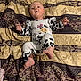 baby, infant, onesie, cow_pattern, blanket, patterned_blanket, bed, foot, hand, face, clothing, cute, lying_down, indoors, child, newborn, soft_lighting, expression, curious, person