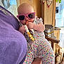 Brooklin joined the competition — help win amazing prizes! baby, sunglasses, pacifier, floral_outfit, infant, indoor, person, hand, wooden_wall, kitchen, chair, boots, purple_shirt, holding, toy, cute, child, casual, home, accessory