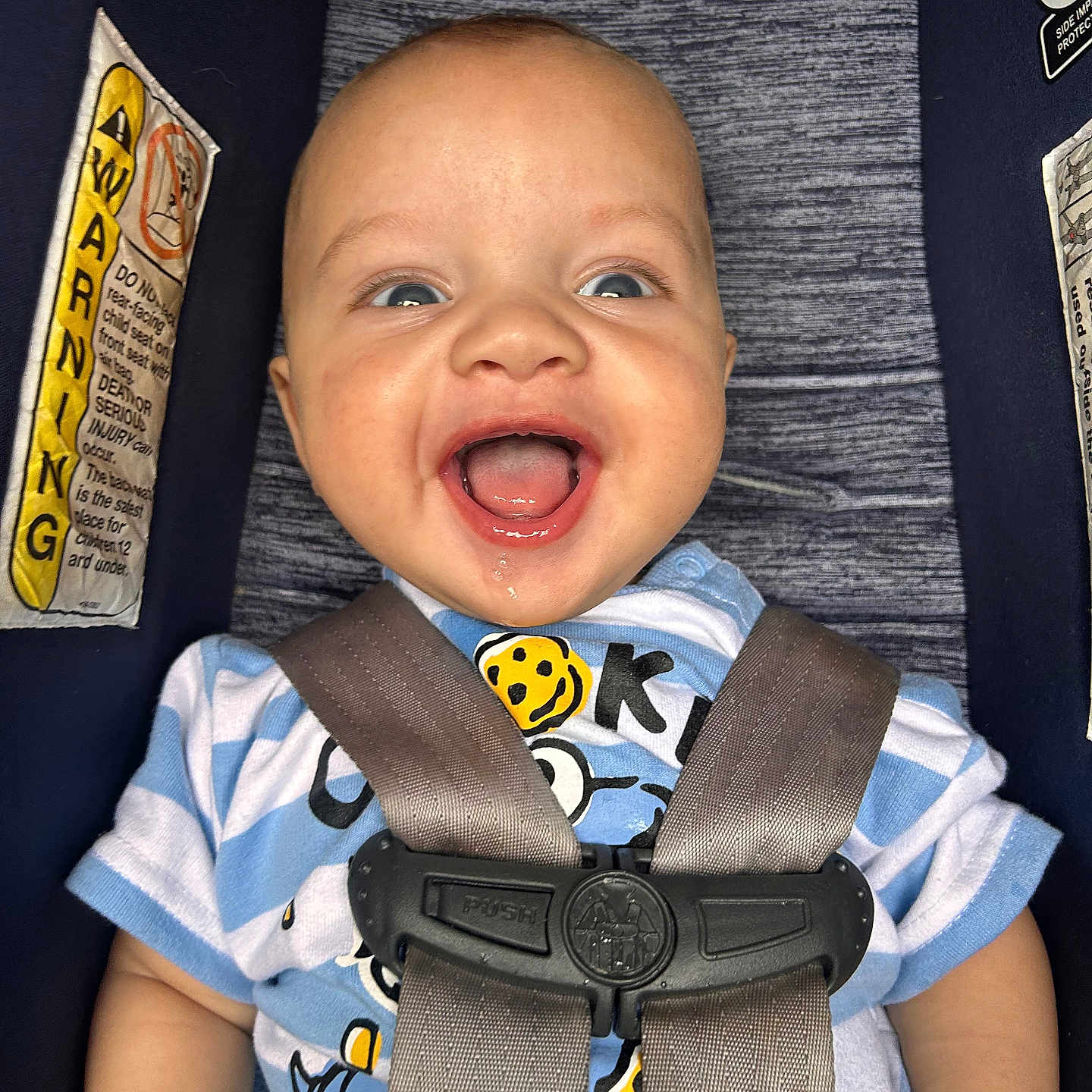 Levi is registered to the contest to win money with this photo: baby, child, car_seat, safety_harness, smiling, happy, infant, blue_shirt, striped_clothing, seat_belt, warning_label, close_up, indoor, cute, face, head, person, sitting, mouth_open, eyes