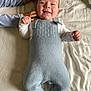 baby, child, smile, knitted_overalls, blue_clothing, white_shirt, hand, blanket, bed, sibling, cute, infant, happy, lying_down, face, person, soft_texture, cozy, indoor, young_child