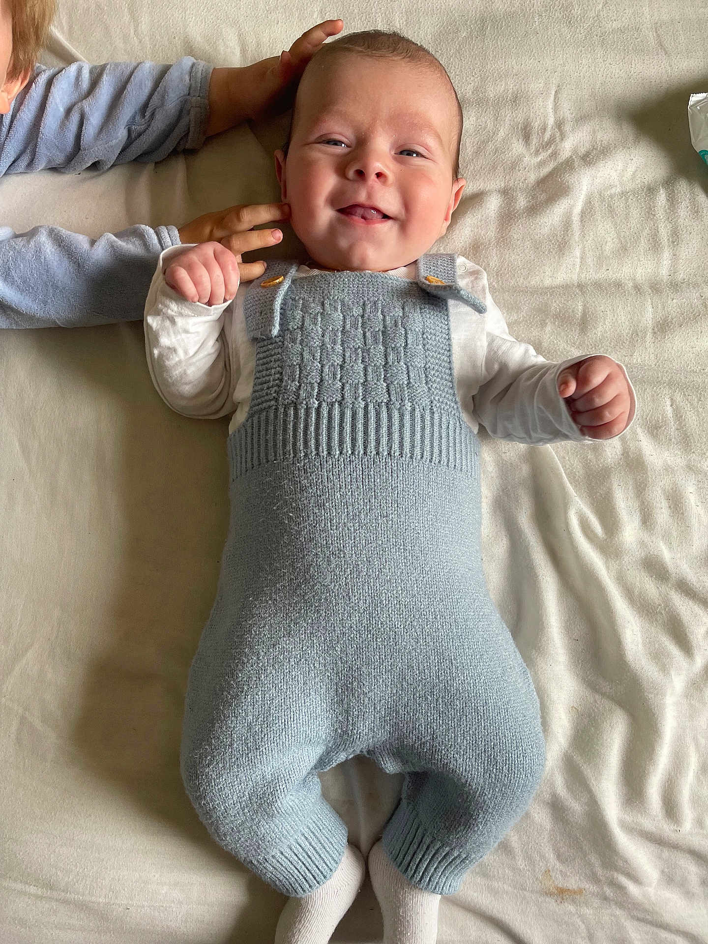 Hugo a rejoint le concours — aidez-le/la à gagner de superbes lots ! baby, child, smile, knitted_overalls, blue_clothing, white_shirt, hand, blanket, bed, sibling, cute, infant, happy, lying_down, face, person, soft_texture, cozy, indoor, young_child
