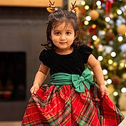 Aanaya joined the competition — help win amazing prizes! child, toddler, girl, holiday_dress, plaid_skirt, green_bow, antler_headband, christmas_tree, ornaments, bokeh_lights, fireplace, festive, portrait, smile, indoors, black_top, red_green, cute, holiday_decor, standing