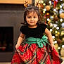 child, toddler, girl, holiday_dress, plaid_skirt, green_bow, antler_headband, christmas_tree, ornaments, bokeh_lights, fireplace, festive, portrait, smile, indoors, black_top, red_green, cute, holiday_decor, standing