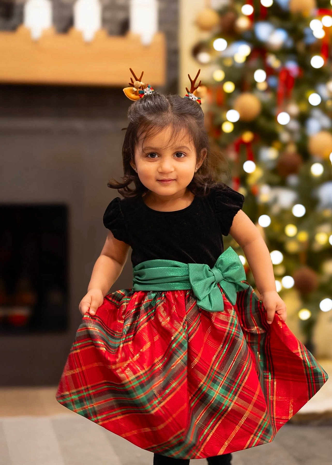 Aanaya joined the competition — help win amazing prizes! child, toddler, girl, holiday_dress, plaid_skirt, green_bow, antler_headband, christmas_tree, ornaments, bokeh_lights, fireplace, festive, portrait, smile, indoors, black_top, red_green, cute, holiday_decor, standing