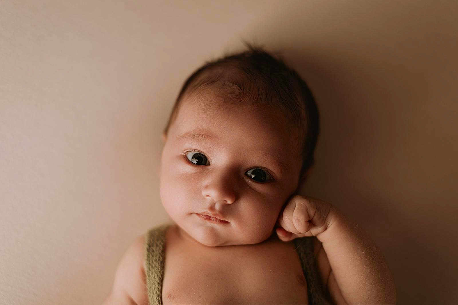 Liam participe au concours pour gagner de l'argent avec cette photo : baby, newborn, infant, portrait, close_up, face, eyes, hand, fist, skin, soft_lighting, beige_background, sleepy, cute, photography, head, clothing, shoulder, expression, studio_shot