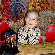 Lény a rejoint le concours — aidez-le/la à gagner de superbes lots ! child, smiling, striped_shirt, lantern, wooden_table, autumn_leaves, pumpkin, hay_bale, wagon_wheel, rustic, cozy, indoors, portrait, face, hands, decor, seasonal, fall, warmth, cute