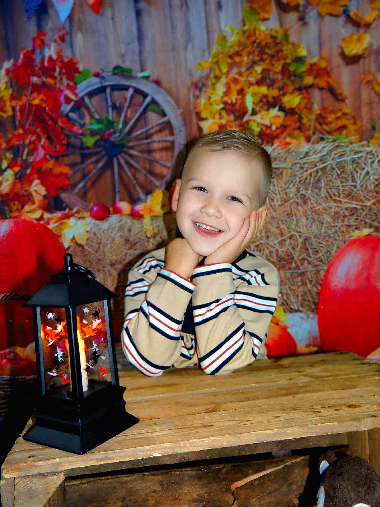 Lény a rejoint le concours — aidez-le/la à gagner de superbes lots ! child, smiling, striped_shirt, lantern, wooden_table, autumn_leaves, pumpkin, hay_bale, wagon_wheel, rustic, cozy, indoors, portrait, face, hands, decor, seasonal, fall, warmth, cute