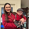 woman, baby, indoor, red_clothing, smile, serious, hat, suspenders, cabinet, furniture, portrait, holding, person, child, family, home, long_hair, fashion, happy, casual
