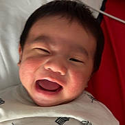 Mung is registered to the contest to win money with this photo: baby, child, smiling, laughing, white_shirt, patterned_clothing, lying_down, rosy_cheeks, face, happy, indoor, pillow, fabric, red, white, cute, infant, person, portrait, joyful