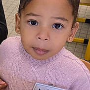 Nila a rejoint le concours — aidez-le/la à gagner de superbes lots ! child, girl, sweater, name_tag, wooden_blocks, table, indoor, curly_hair, bun, pink, play, toy, education, classroom, seat, person, face, young, cute, learning