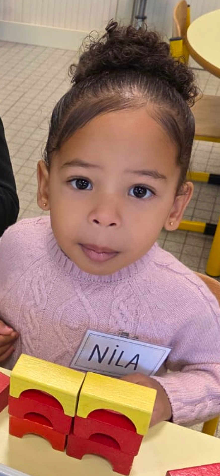 Nila a rejoint le concours — aidez-le/la à gagner de superbes lots ! child, girl, sweater, name_tag, wooden_blocks, table, indoor, curly_hair, bun, pink, play, toy, education, classroom, seat, person, face, young, cute, learning