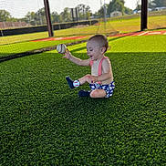Weston is registered to the contest to win money with this photo: baby, child, baseball, sportswear, grass, turf, outdoor, playing, happy, sitting, sunlight, smiling, socks, suspenders, field, netting, daytime, recreation, fun, joyful