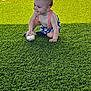 Weston is registered to the contest to win money with this photo: baby, child, baseball, grass, artificial_turf, sports_field, suspender, shorts, smiling, crouching, outdoor, sunlight, net, running_track, green, red, playing, person, happy, summer