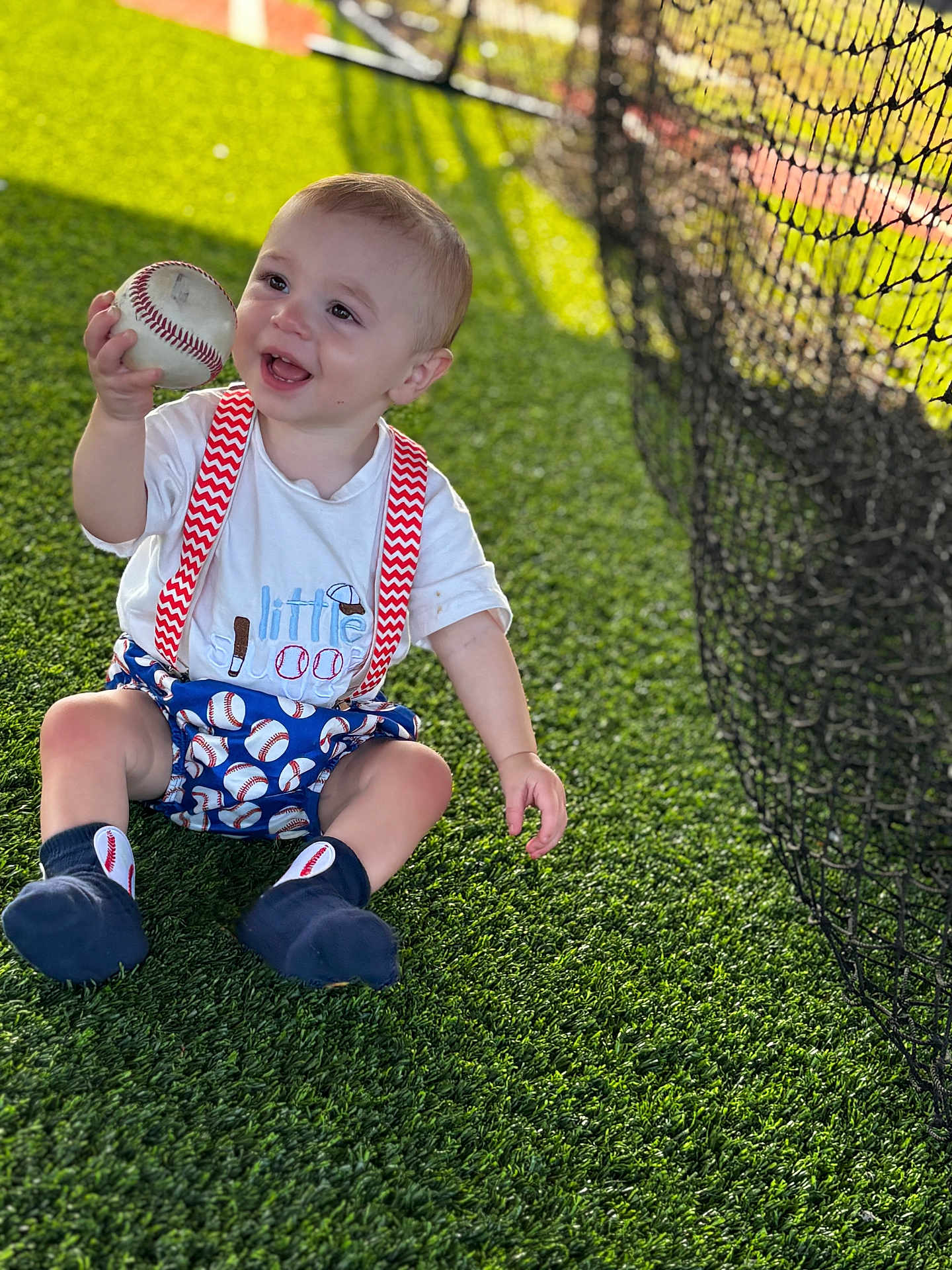Weston is registered to the contest to win money with this photo: toddler, child, baseball, grass, artificial_turf, net, fence, outdoor, sunlight, happy, smiling, sitting, clothing, socks, suspenders, play, sport, ball, person, cute