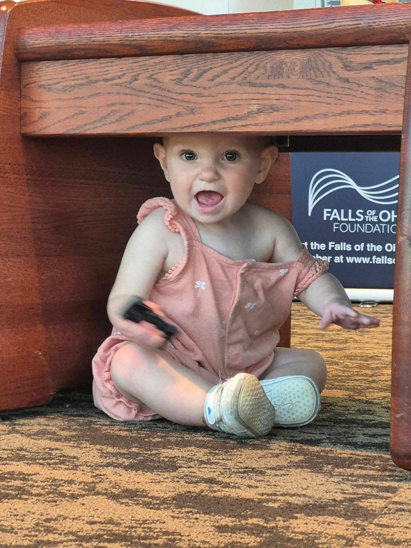 Ahna joined the competition — help win amazing prizes! baby, child, romper, sitting, carpet, wooden_table, indoor, happy, curious, person, footwear, shoe, smile, expression, playful, small_object, furniture, floor, candid, young