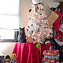 cat, christmas_tree, holiday_decorations, presents, stockings, snowman, ornaments, table, red_tablecloth, window, lamp, indoor, festive, decor, wrapped_gifts, holiday, smiling_snowman, gray_cat, christmas, cozy