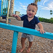 Atlas joined the competition — help win amazing prizes! toddler, child, playground, barefoot, smiling, curly_hair, blue_shirt, shorts, metal_bar, wood_chips, outdoor, daylight, palm_trees, sky, fence, park, happy, person, play, summer