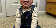 Octavia is registered to the contest to win money with this photo: baby, infant, toddler, smile, tongue_out, denim_overalls, overalls, socks, paw_prints, carpet, sitting, hands, door, dresser, indoor, portrait, blurred_background, happy, cute, footwear