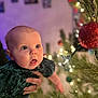 baby, infant, child, christmas_tree, ornament, christmas_lights, bokeh, surprised_expression, person, hand, green_dress, festive, holiday, portrait, indoor, closeup, blue_eyes, rosy_cheeks, parent_hand, decoration