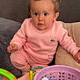 toddler, child, pink_outfit, pigtails, couch, toy_basket, colorful_toys, cushions, indoor, curious, playing, seated, young_child, casual_clothing, home_environment, soft_lighting, portrait, person, baby_toys, relaxed