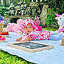 baby, floral_hat, floral_dress, blanket, grass, cupcakes, strawberries, oranges, stuffed_animals, unicorn, pink_teddy_bear, basket, outdoor, garden, stone_wall, greenery, child, picnic, toy, cute