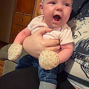 Hadley is registered to the contest to win money with this photo: baby, infant, person, adult_hand, headband, bow, mittens, sock, onesie, jeans, sitting, lap, couch, wood_paneling, cabinet, drawer, indoor, happy, open_mouth, blurred